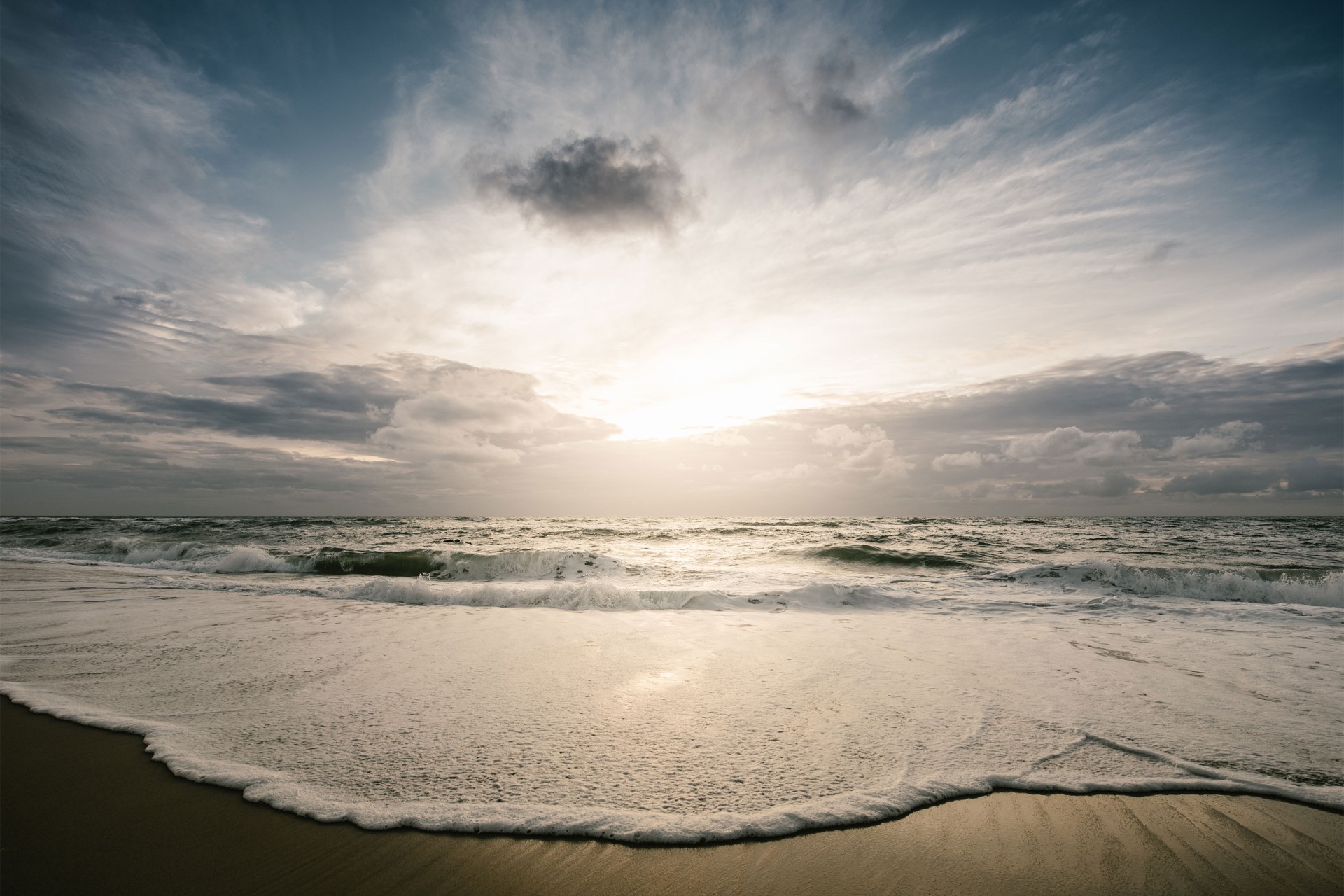 Eine Welle läuft auf den Strand, dahinter unruhiges Wasser und ein Sonnenuntergang.