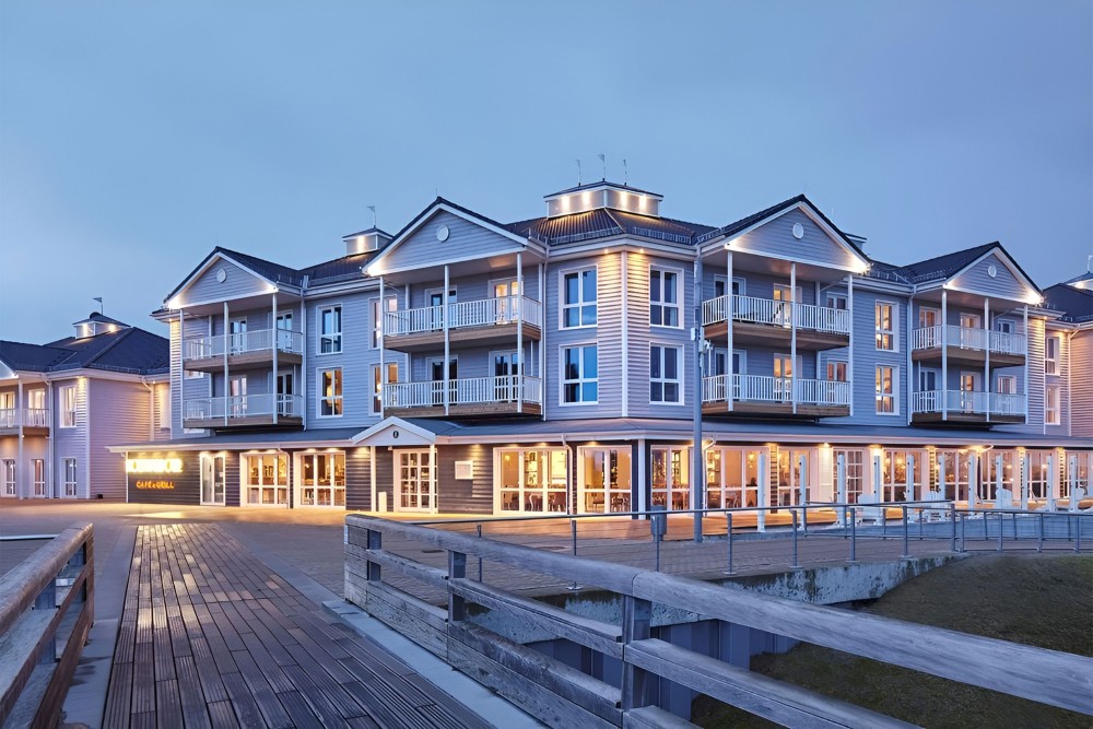 Blick von einer Holzterasse auf ein Hotel mit Balkonen in abendlicher Stimmung, dahinter blauer Himmel.