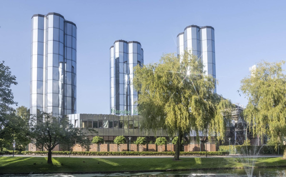 Drei hohe, zylindrische Glasgebäude stehen neben einem Teich mit Springbrunnen und Bäumen.