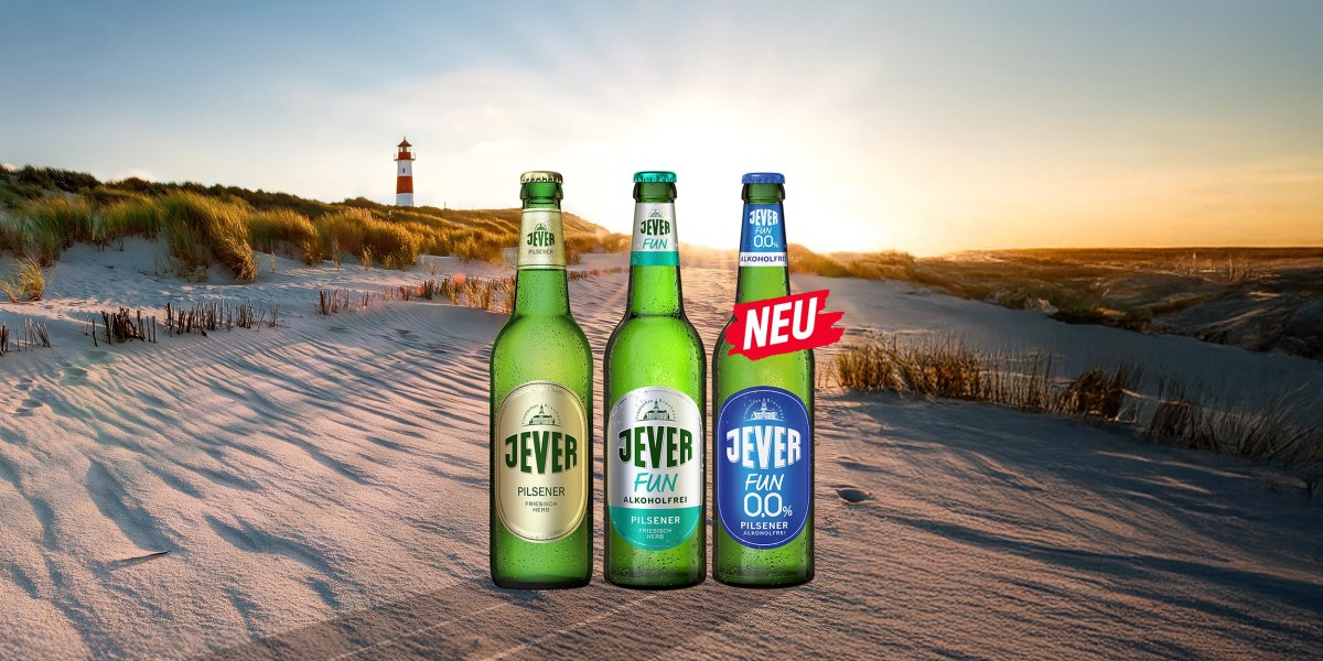 3 Bierflaschen der Sorten Jever, Jever Fun und Jever Fun 0,0% stehen im Sand am Strand, im Hintergrund ein Leuchtturm mit Sonnenuntergang.