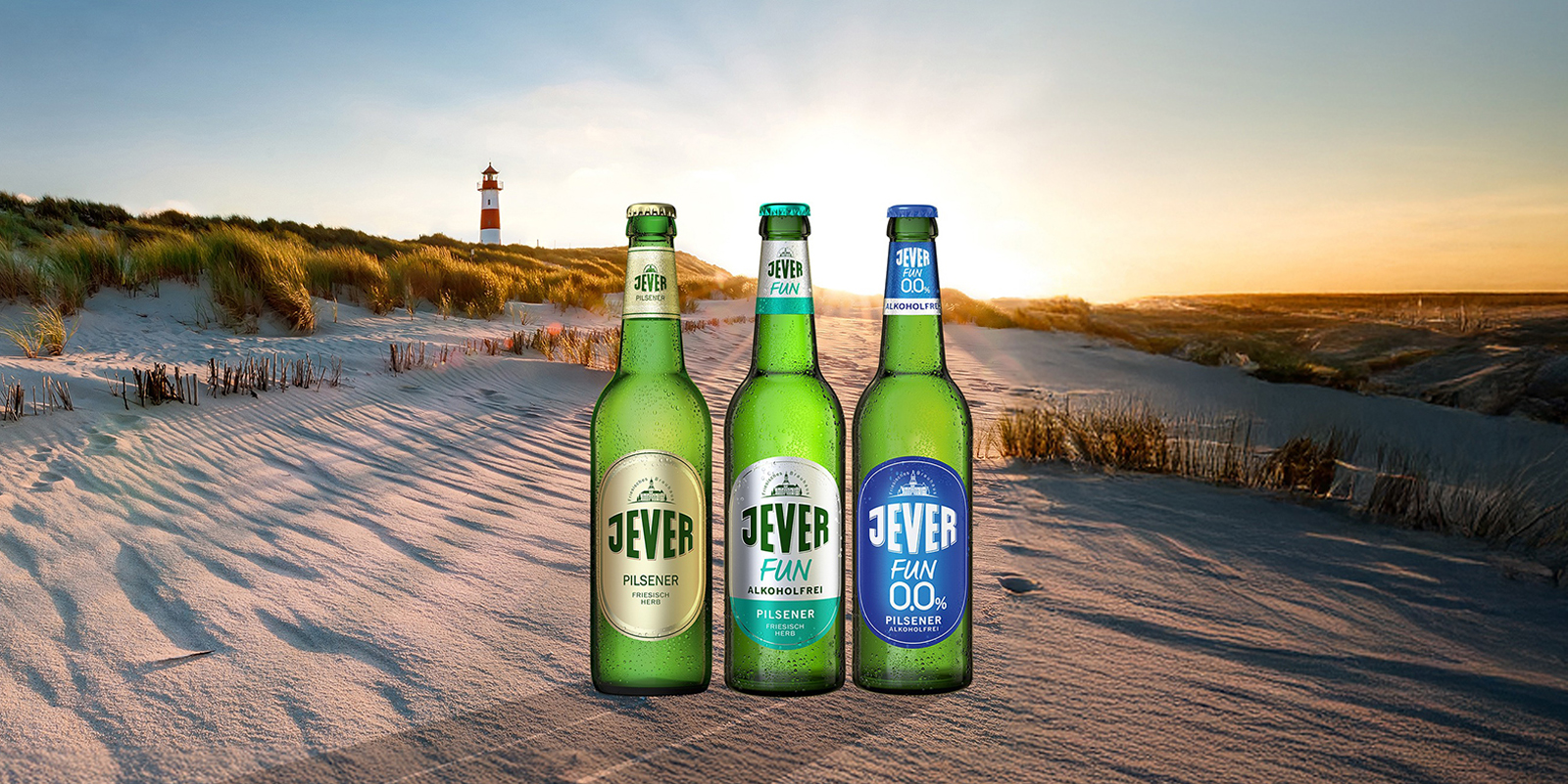 3 Bierflaschen der Sorten Jever, Jever Fun und Jever Fun 0,0 % stehen im Sand am Strand, im Hintergrund ein Leuchtturm mit Sonnenuntergang.