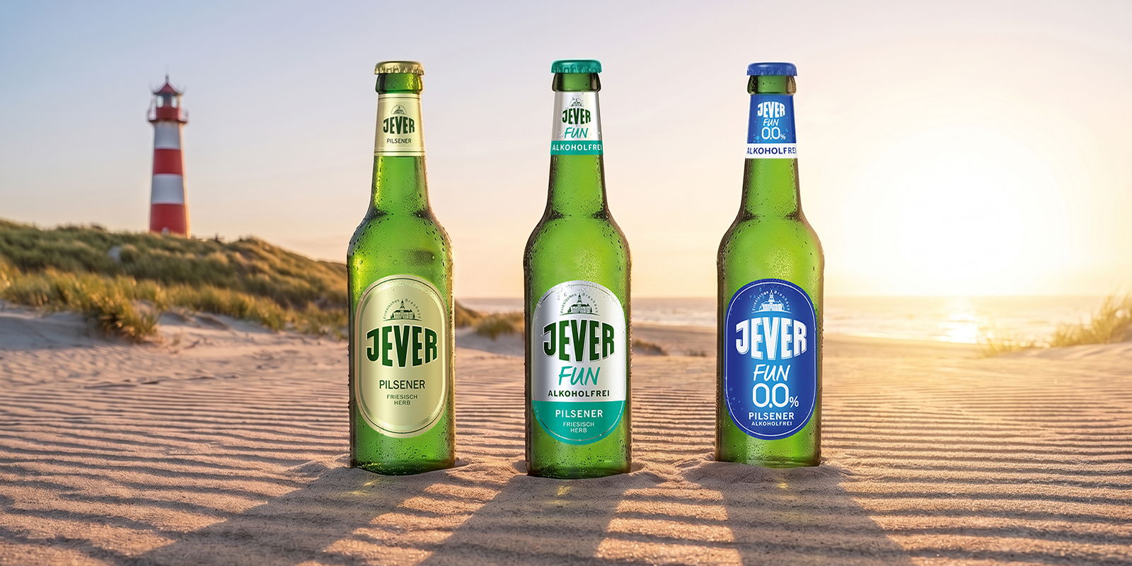 3 Bierflaschen der Sorten Jever, Jever Fun und Jever Fun 0,0% stehen im Sand am Strand, im Hintergrund ein Leuchtturm mit Sonnenuntergang.