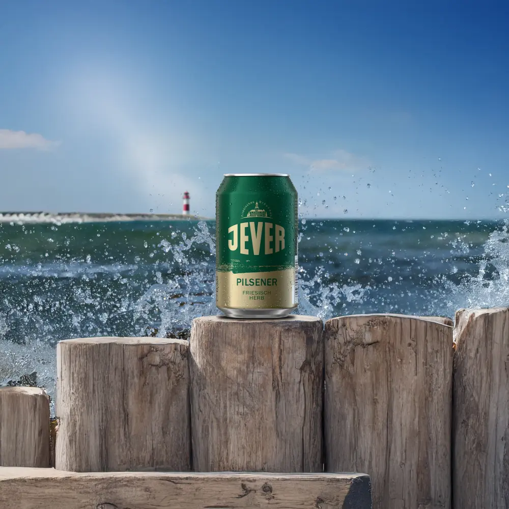 Grüne Jever-Dose steht auf Holzpfählen am Meer mit Gischt. Im Hintergrund Wellen, blauer Himmel und ein Leuchtturm.