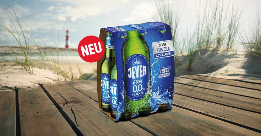 Ein Jever Fun 0,0 % Sixpack steht auf einem Strandsteg, dahinter das Meer und ein Leuchtturm.