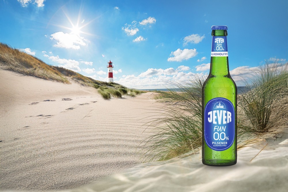 Ein Leuchtturm steht auf einer Düne am Strand unter einem blauen Himmel mit Sonne, im Vordergrund steht ein Flasche Jever Fun 0,0 Prozent Alkohol.