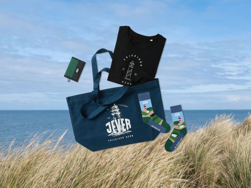 Verschiedene Jever Werbemittel liegen auf einem Strandgras bedeckten Sanddüne, dahinter das Meer.